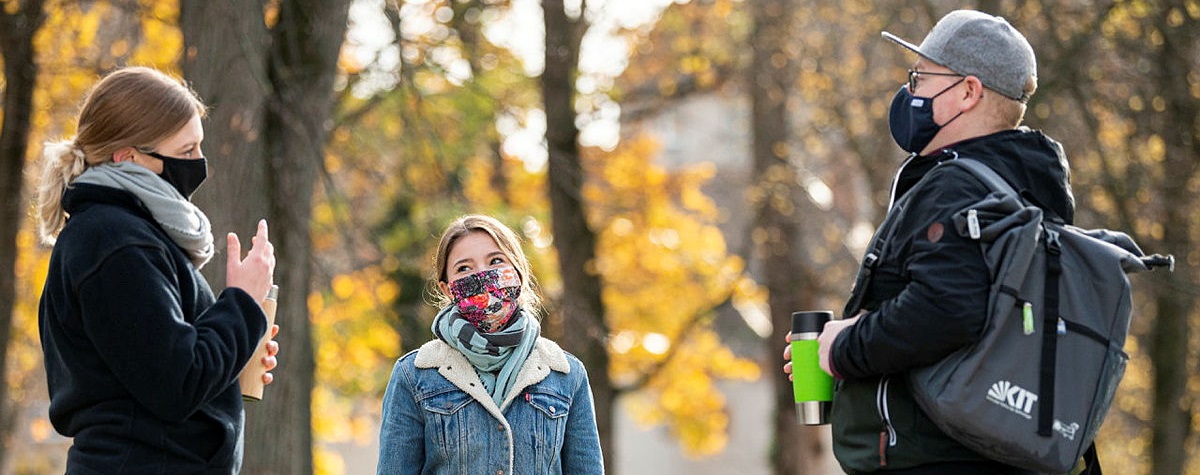 Drei Studierende mit Masken draußen im Gespräch. Herbstlicher Hintergrund, Bäume, gorldene Blätter.